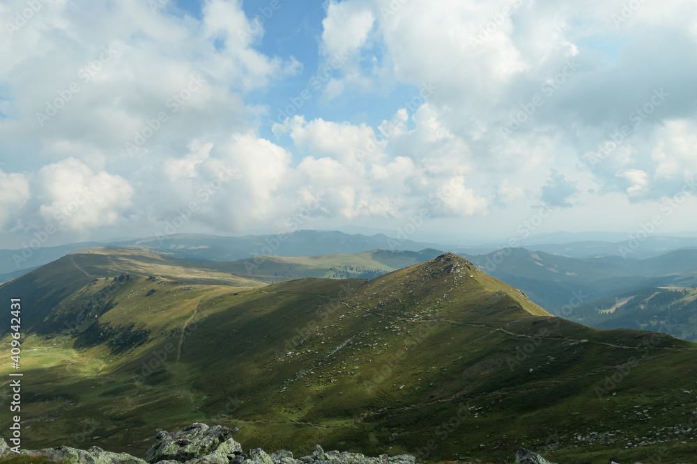 A panoramic view on the distant Grosse Speikkogel in Austrian Alps. The mountains around are lush green. A bit of overcast, blue sky. Soft and edgeless mountains. Achievement and completion.