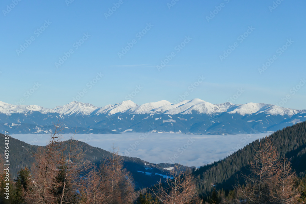 An early, wintery morning in the region of Stubalpe, Austria. A thick fog in the valley below. The distant mountain peaks are covered with powder snow. Dried trees on the slopes. Day break. An early, wintery morning in the region of Stubalpe, Austria. A thick fog in the valley below. The distant mountain peaks are covered with powder snow. Dried trees on the slopes. Day break.
