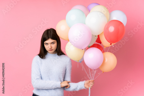 Young Ukrainian teenager girl holding lots of balloons over isolated pink background keeping arms crossed