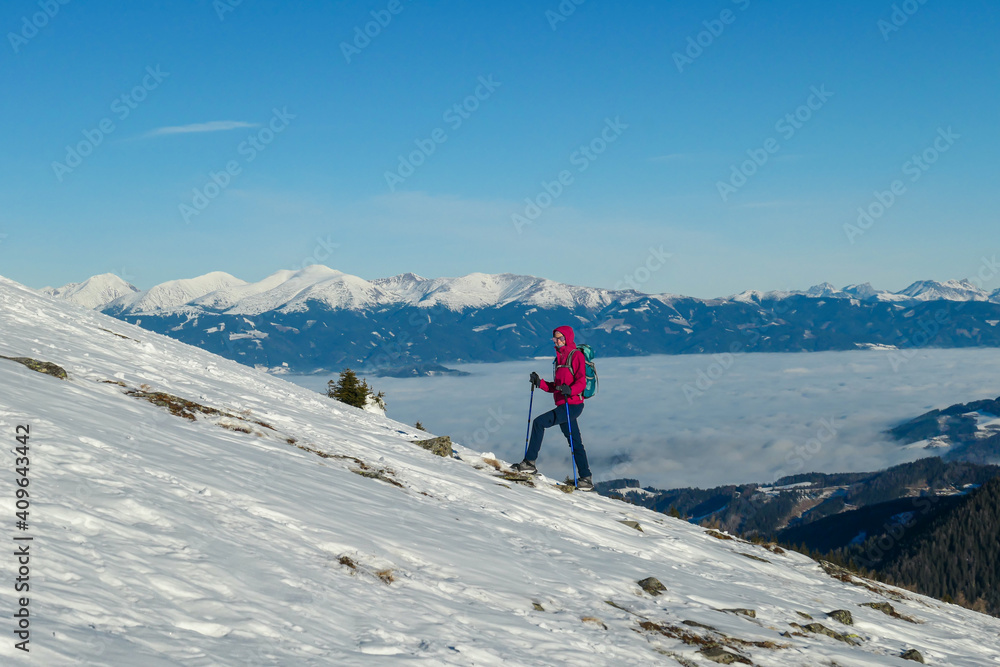 A woman wearing pink jacket and snow shoes hiking up to Amerinkogel's peak in Austrian Alps. Fresh powder snow. Many mountain chains in the back, valley shrouded in fog. Winter outdoor activity. Fun