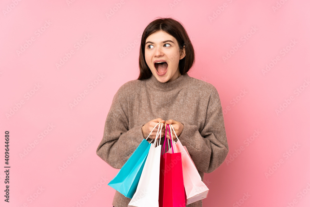 Young Ukrainian teenager girl over isolated pink background holding shopping bags and surprised
