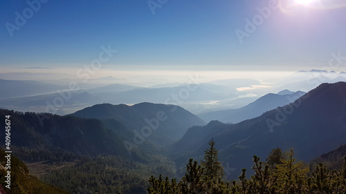 Fototapeta Naklejka Na Ścianę i Meble -  A panoramic view on the haze shrouded valley from the way to Mittagskogel in Austrian Alps. Clear and sunny day. Endless mountain chains. Outdoor activity. There are a few pine trees on the bottom