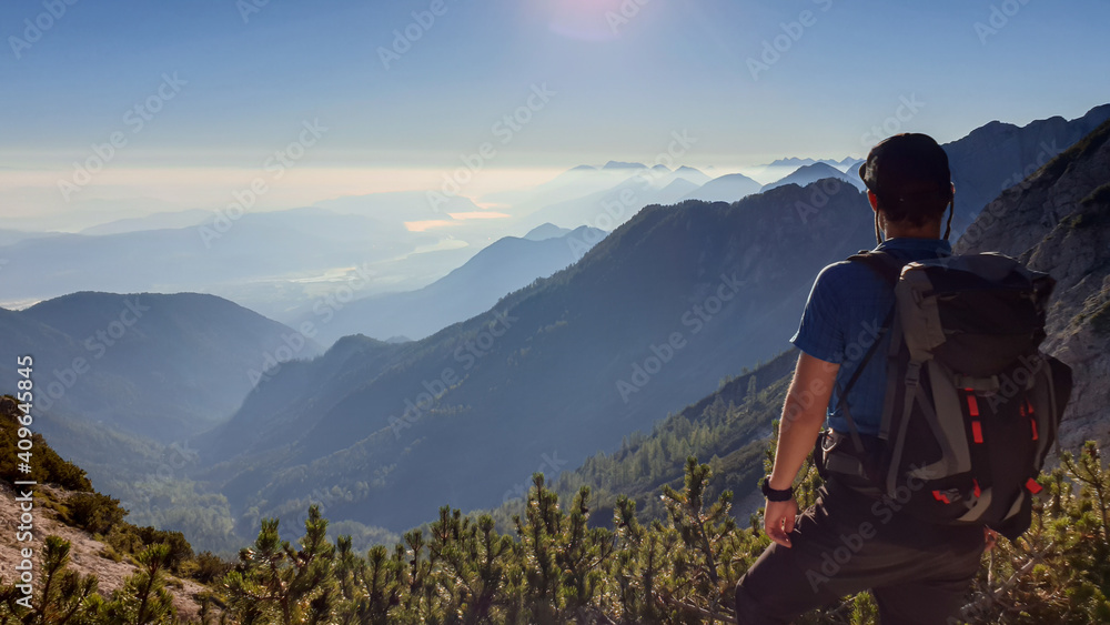 Fototapeta premium A man in a hiking outfit with a panoramic view on the haze shrouded valley from the way to Mittagskogel in Austrian Alps. Clear and sunny day. Endless mountain chains. Outdoor activity. Achievement