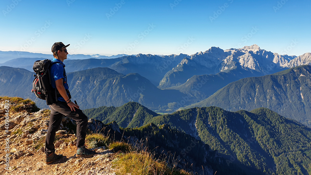 Fototapeta premium A man in a hiking outfit with a panoramic view on the haze shrouded valley from the way to Mittagskogel in Austrian Alps. Clear and sunny day. Endless mountain chains. Outdoor activity. Achievement