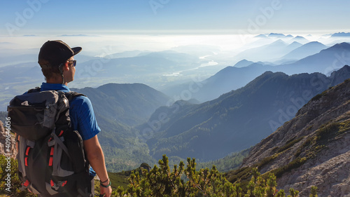 A man in a hiking outfit with a panoramic view on the haze shrouded valley from the way to Mittagskogel in Austrian Alps. Clear and sunny day. Endless mountain chains. Outdoor activity. Achievement