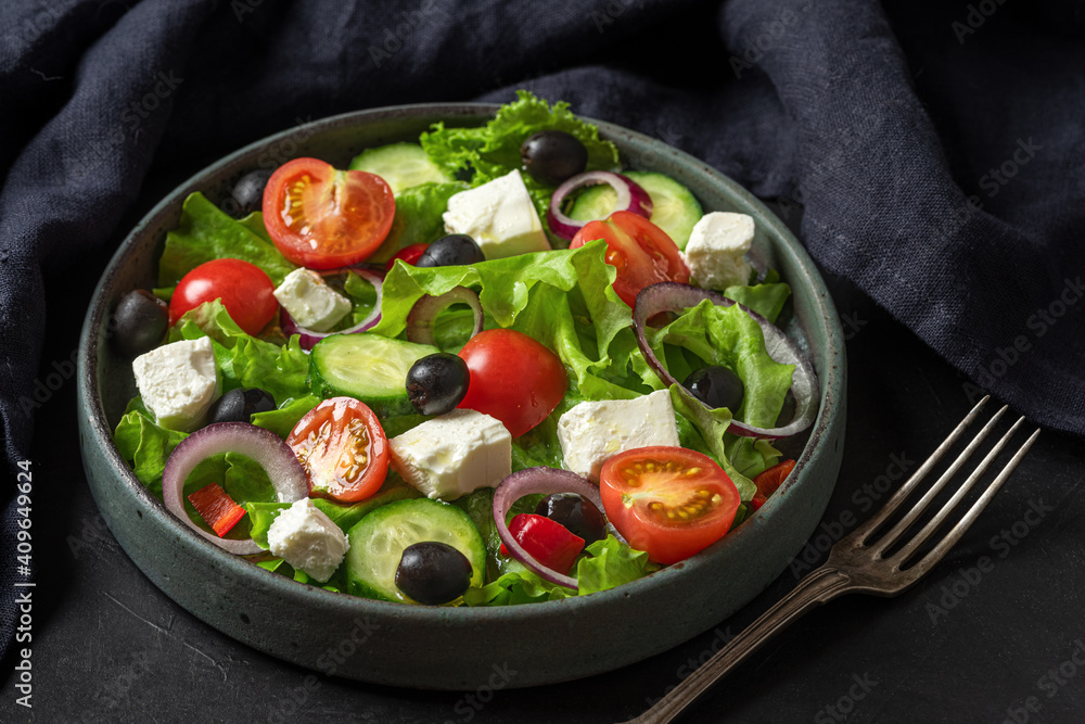 Greek salad with fresh vegetables, feta cheese and olives in a plate with fork and towel on black background
