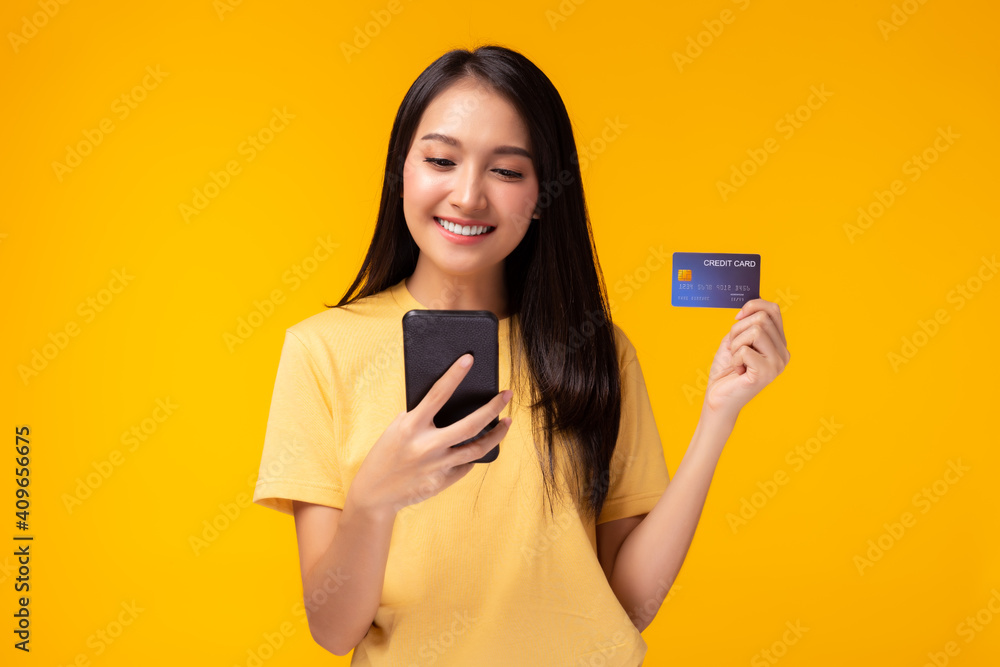 Portrait happy young asian girl standing over yellow background, holding mobile phone, showing credit card. Online payment, hands holding credit card, using smart phone for online shopping Portrait happy young asian girl standing over yellow background, holding mobile phone, showing credit card. Online payment, hands holding credit card, using smart phone for online shopping