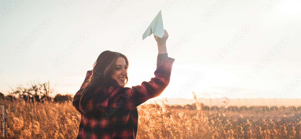 Travelling woman with paper airplane enjoying life and freedom at the land at sunset. Arms outstretched and happiness - obrazy, fototapety, plakaty 