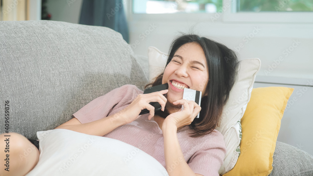 Young smiling Asian woman using smartphone buying online shopping by credit card while lying on sofa when relax in living room at home. Lifestyle latin and hispanic ethnicity women at house concept. Young smiling Asian woman using smartphone buying online shopping by credit card while lying on sofa when relax in living room at home. Lifestyle latin and hispanic ethnicity women at house concept.
