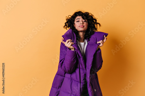 Lovable young woman posing in purple down jacket. Front view of glamorous curly mixed race girl isolated on yellow background. © Look!