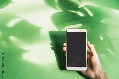 Hand holding empty screen smart phone with shadow of tropical green leave on color background with copy space, Top view
