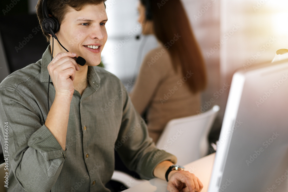 Young dark-haired guy with headsets is talking to a client, while sitting in sunny office. Call center operators at work Young dark-haired guy with headsets is talking to a client, while sitting in sunny office. Call center operators at work