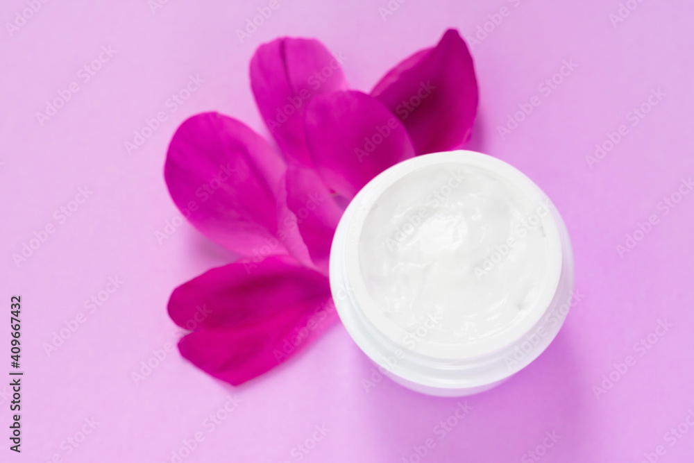 Container full of white facial cream with peony petals nearby on pink background Container full of white facial cream with peony petals nearby on pink background