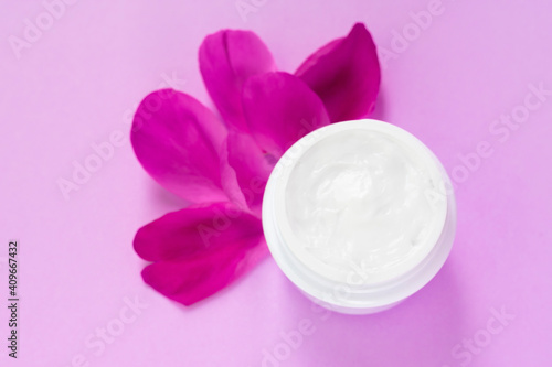Container full of white facial cream with peony petals nearby on pink background