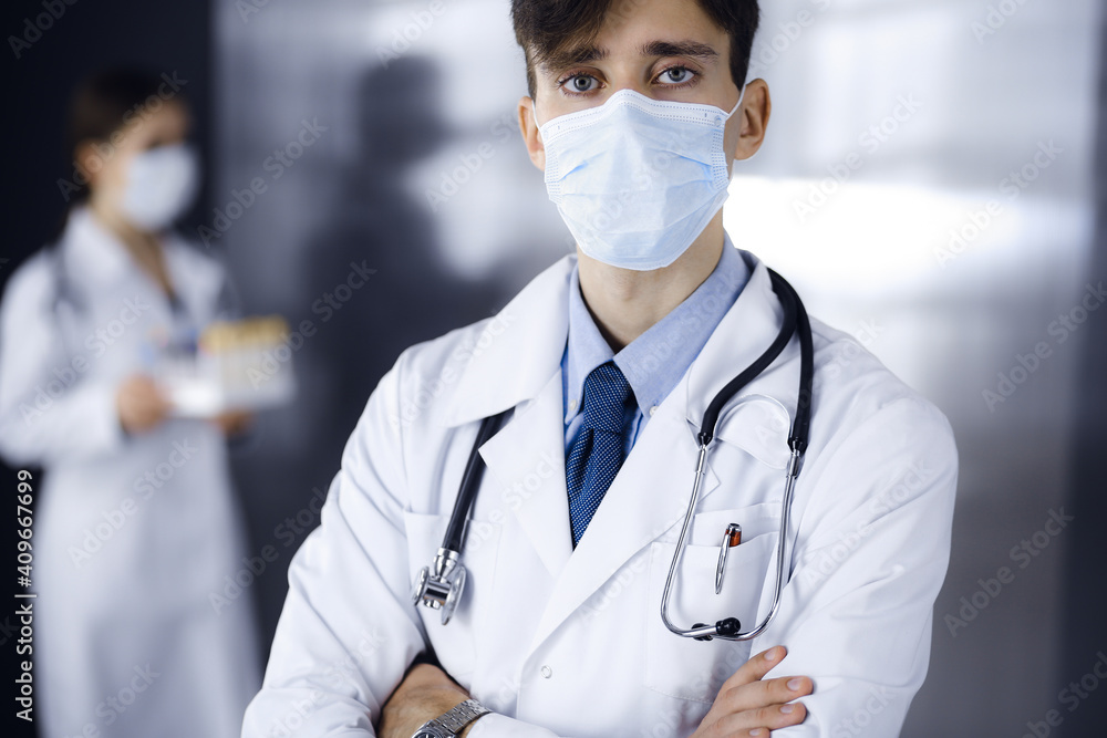 Young male doctor standing with arms crossed in clinic. Smart physician wearing medical protective mask in modern hospital. Medicine and Corona virus concept Young male doctor standing with arms crossed in clinic. Smart physician wearing medical protective mask in modern hospital. Medicine and Corona virus concept