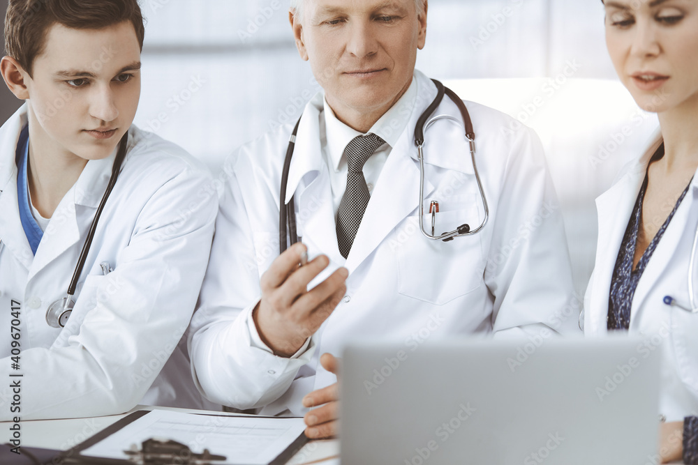 Doctors colleagues and elderly therapist discussing current disease therapy while sitting at working place in sunny clinic. Teamwork in medicine Doctors colleagues and elderly therapist discussing current disease therapy while sitting at working place in sunny clinic. Teamwork in medicine