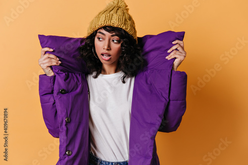 Sensual young woman in purple down jacket looking away. Studio shot of cute mixed race girl in knitted hat. © Look!