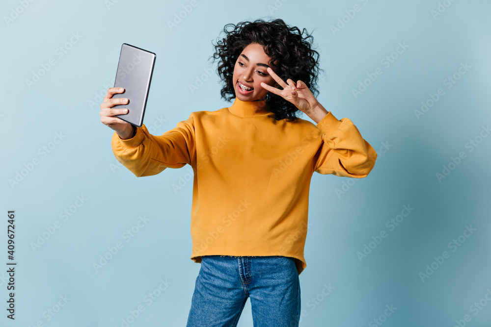 Mixed race girl posing with peace sign. Carefree young woman using digital tablet on blue background. Mixed race girl posing with peace sign. Carefree young woman using digital tablet on blue background.