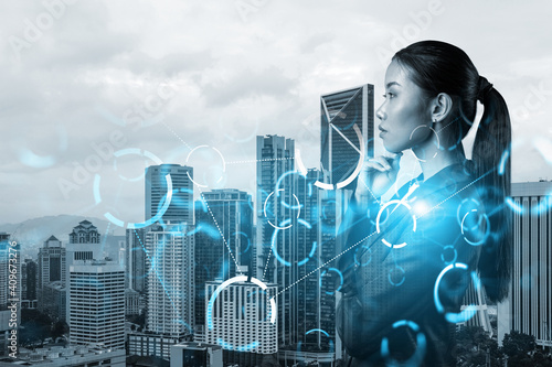 Attractive young Asian businesswoman pondering on technology at business process to achieve tremendous growth in commerce. Tech hologram icons over Kuala Lumpur background