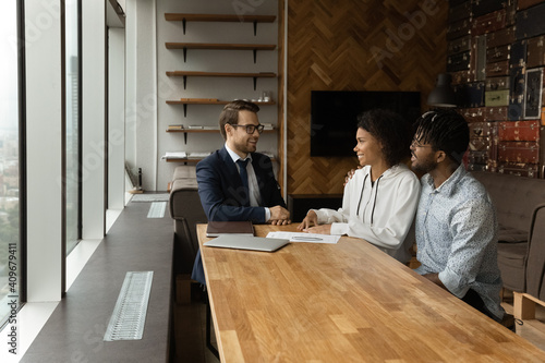 Happy african american couple sit by desk at travel agent office listen to pleasant male consultant choose trip. Confident young businessman talk with black husband wife customers offer house to buy photo