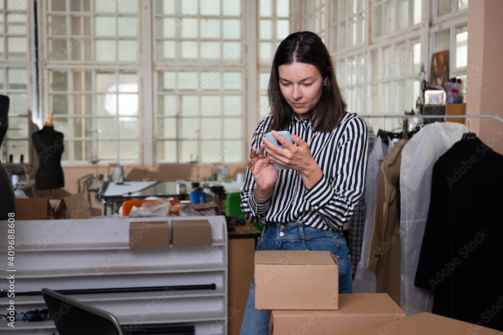 Small business, tailor shop, costume designer's workshop. A young woman entrepreneur in the workplace is the packaging of the parcel and uses the phone for business contacts. Portrait in the interior