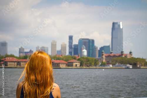 A young woman with long red hair is watching the Manhattan skyline from Liberty Island in New York City