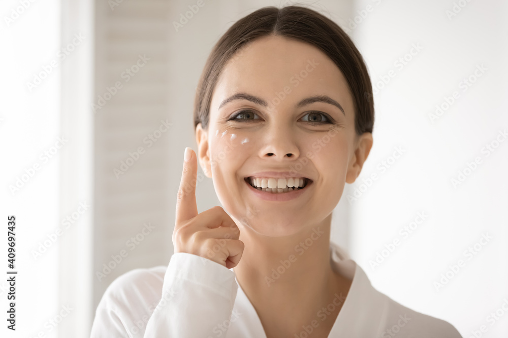 Headshot portrait of smiling young Caucasian woman in bathrobe do daily beauty treatment or procedures at home bathroom. Happy millennial female apply nourishing moisturizing face cream on skin. Headshot portrait of smiling young Caucasian woman in bathrobe do daily beauty treatment or procedures at home bathroom. Happy millennial female apply nourishing moisturizing face cream on skin.