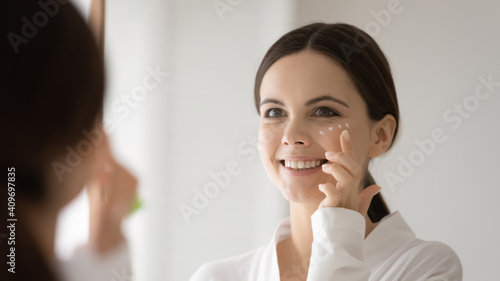 Smiling Caucasian woman look in mirror apply moisturizing facial cream for soft healthy skin. Happy young female use nourishing balm or serum, do daily facial beauty procedures. Skincare concept.
