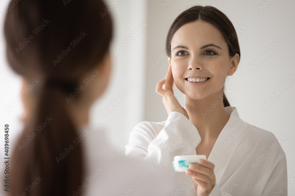 Smiling young Caucasian woman look in mirror hold bottle apply nourishing face cream for healthy soft skin. Happy female do morning beauty procedures with cosmetic product. Skincare concept. Smiling young Caucasian woman look in mirror hold bottle apply nourishing face cream for healthy soft skin. Happy female do morning beauty procedures with cosmetic product. Skincare concept.