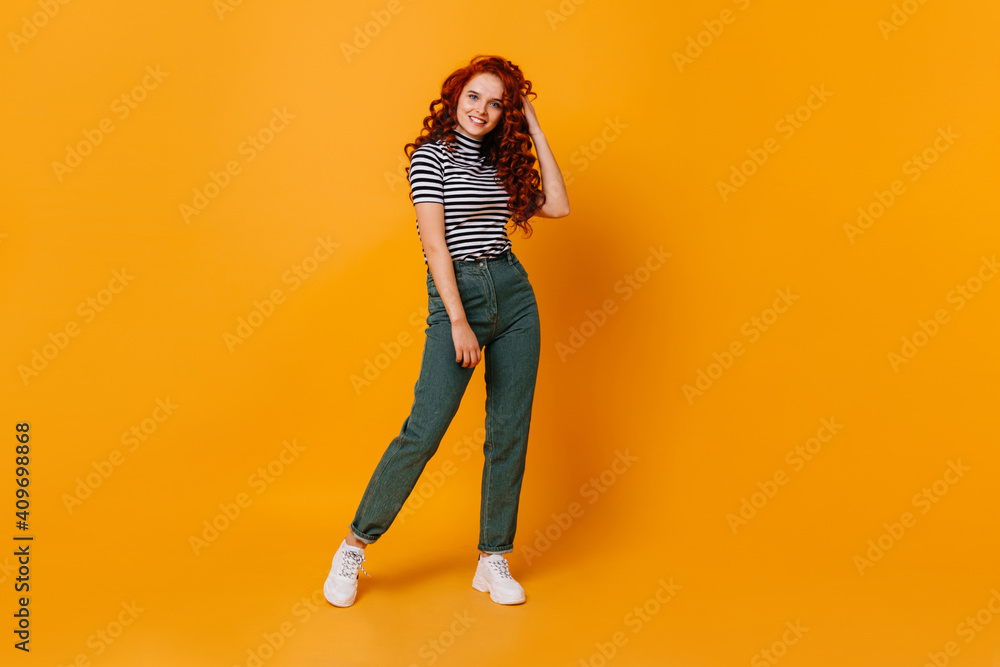 Pretty young woman touches her curly red hair and smiles. Portrait of girl in jeans and top in orange studio Pretty young woman touches her curly red hair and smiles. Portrait of girl in jeans and top in orange studio