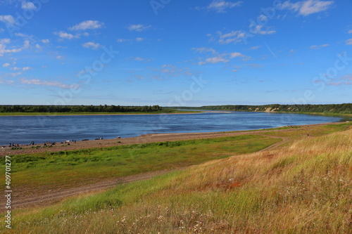 Panoramic image from the coast to open spaces with a wide and long river, meadows and people tourists with boats near the water in the distance on a sunny day with a blue sky.View photography