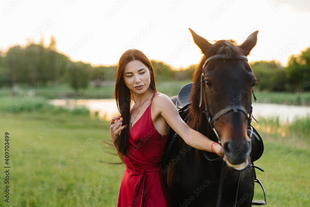 Obraz premium A young pretty girl in a red dress poses on a ranch with a thoroughbred stallion at sunset. Love and care for animals
