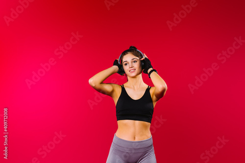 Caucasian sporty girl in a black sporty top and big headphones looks to the side on a red background with empty side space.