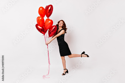 Full length shot of ginger woman in black dress. Studio photo of slim girl dancing with heart shaped balloons. photo