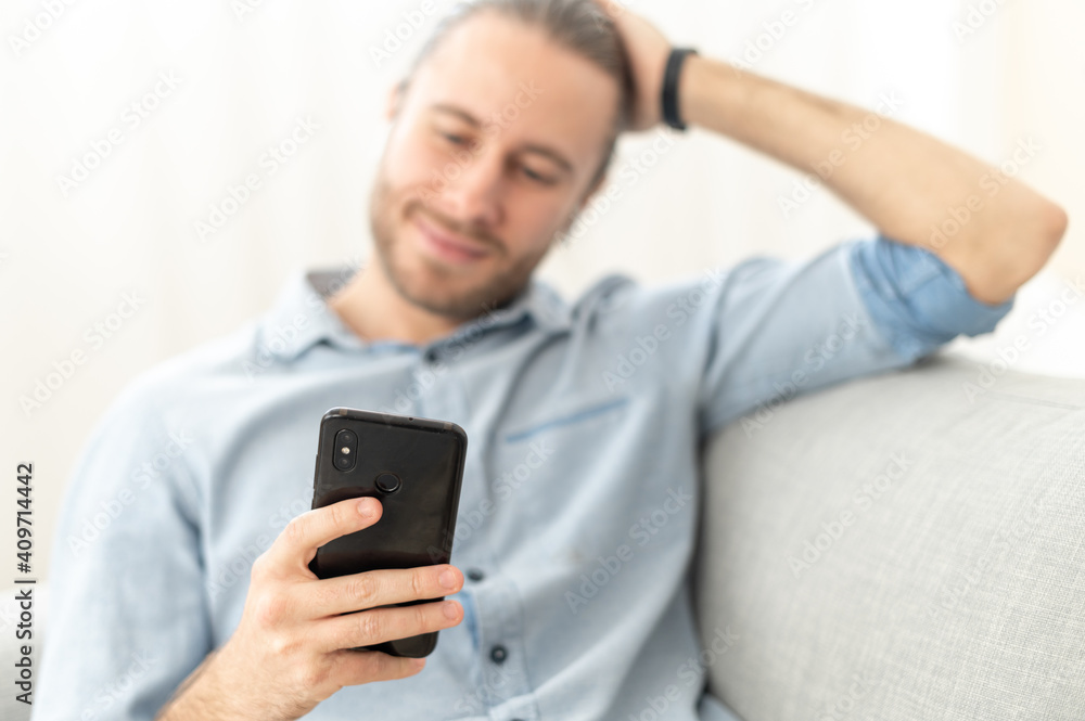 Attractive millennial young bearded hipster man relaxing and sitting on the comfy sofa in the living room, chatting with a friend in social media on mobile phone, dating, spending leisure time online