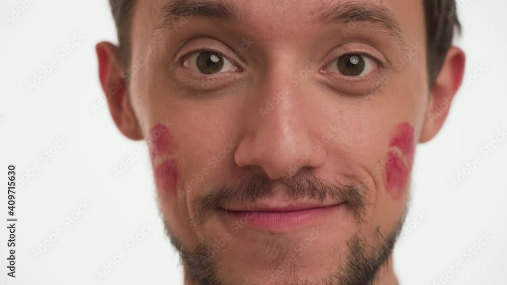 One young attractive brunette man 20s with dark beard, moustache, brown eyes, red kiss imprints or marks on face show spots on cheek, smile isolated on white wall background close up. Fun love concept One young attractive brunette man 20s with dark beard, moustache, brown eyes, red kiss imprints or marks on face show spots on cheek, smile isolated on white wall background close up. Fun love concept