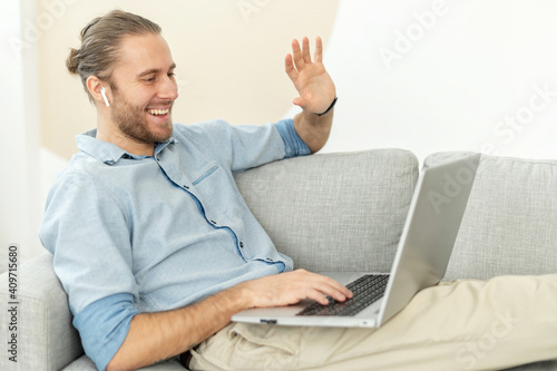 A smiling bearded hipster man is sitting on the comfy couch in the living room, relaxing and talking to his friend or family, waving at the laptop screen, staying connected during pandemic