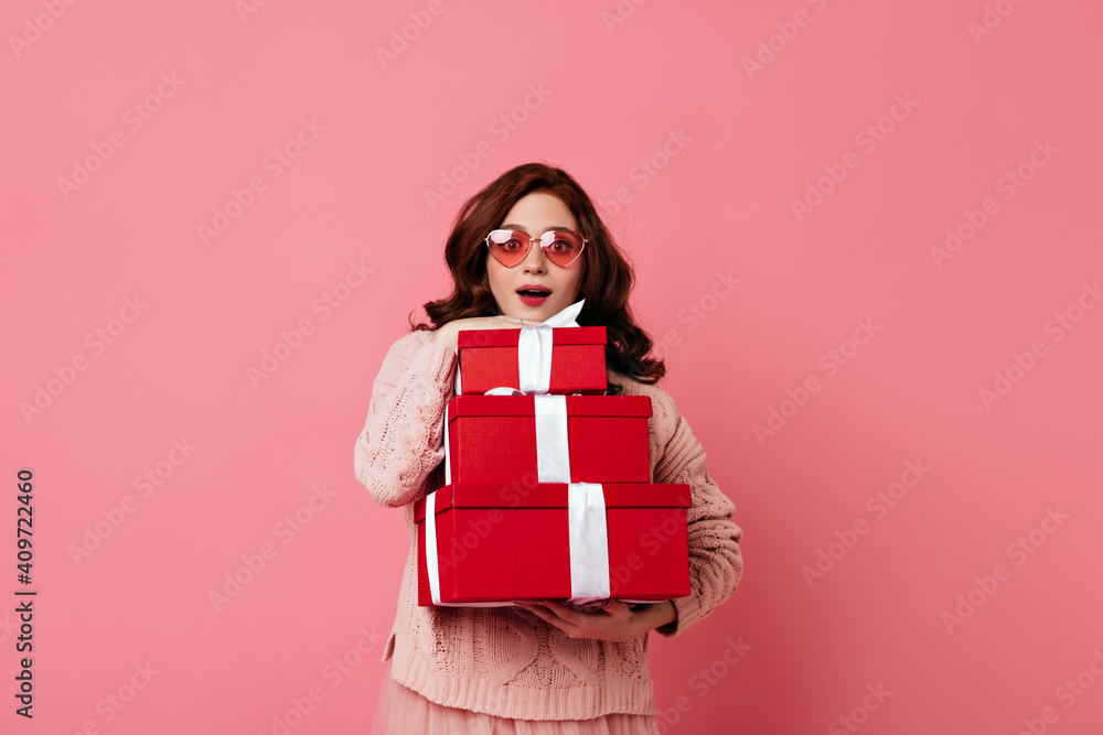Fototapeta premium Amazed ginger lady holding new year presents on pink background. Studio photo of good-looking girl with gifts.