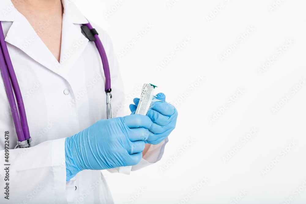 A young nurse in a mask opens a syringe on a white background in a white coat, to make a vaccination, an injection. The concept of vaccination and protection against influenza, from avid, copy space A young nurse in a mask opens a syringe on a white background in a white coat, to make a vaccination, an injection. The concept of vaccination and protection against influenza, from avid, copy space