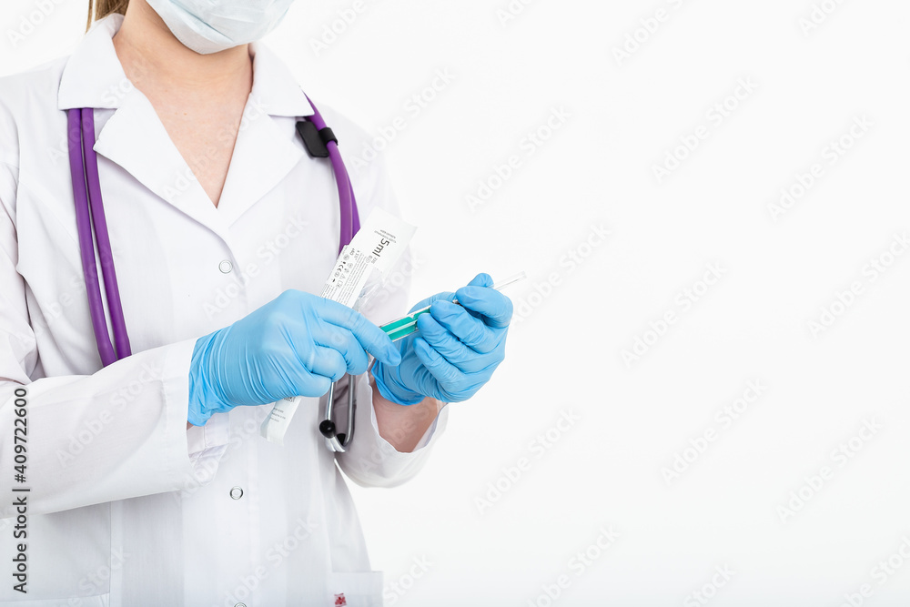 A young nurse in a mask opens a syringe on a white background in a white coat, to make a vaccination, an injection. The concept of vaccination and protection against influenza, from avid, copy space A young nurse in a mask opens a syringe on a white background in a white coat, to make a vaccination, an injection. The concept of vaccination and protection against influenza, from avid, copy space