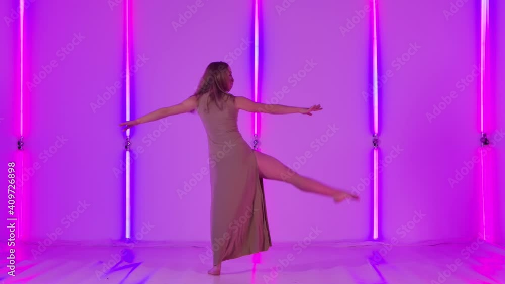 Feminine female dancer performs sensual contempt and jumps gracefully against a backdrop of bright neon lights. Young woman dancing modern choreography in a dark studio. Slow motion. Feminine female dancer performs sensual contempt and jumps gracefully against a backdrop of bright neon lights. Young woman dancing modern choreography in a dark studio. Slow motion.