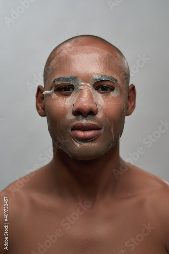 Closeup portrait of young african american man with textile sheet mask applied on his face looking at camera, posing isolated over gray background