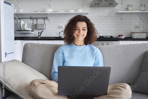 Happy hispanic teen girl holding laptop computer device technology sitting on couch at home. Smiling young woman using apps, searching online, e learning, studying browsing internet on sofa.