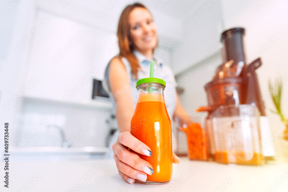 Smiling fit woman making fresh carrot juice in a juicer in the kitchen at home. Bottle of fresh homemade carrot juice on the table at morning sunlight. Healthy lifestyle Smiling fit woman making fresh carrot juice in a juicer in the kitchen at home. Bottle of fresh homemade carrot juice on the table at morning sunlight. Healthy lifestyle