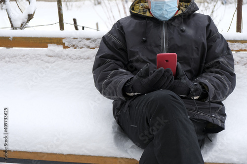 An aged man sitting on a bench covered with snow. Protective mask. Warm mittens. Red phone in men's hands. The man keeps calm. The photo looks like a meme. Copy space.