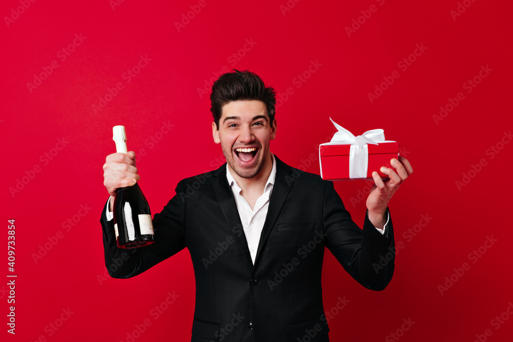 Happy man with wine and present box smiling at camera. Front view of brunette boy with gift isolated on red.