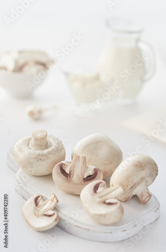 Champignons on a white wooden board in the foreground. In the background, in blur, a bowl of chopped mushrooms and a sauce bowl.