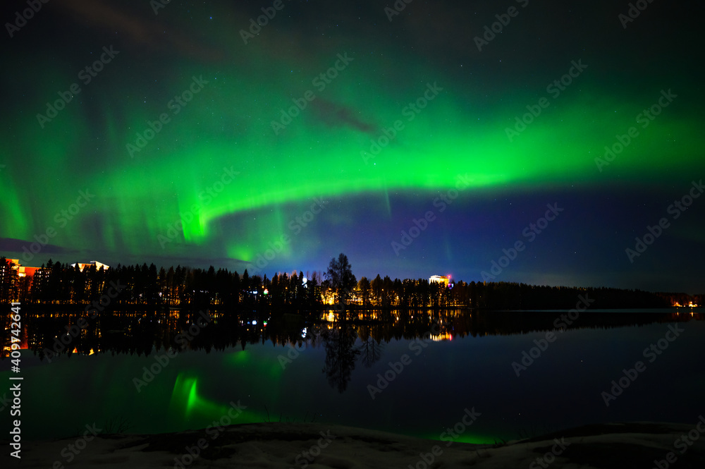 A landscape view of aurora borealis with a city scape under starry clear sky and reflection from lake water
