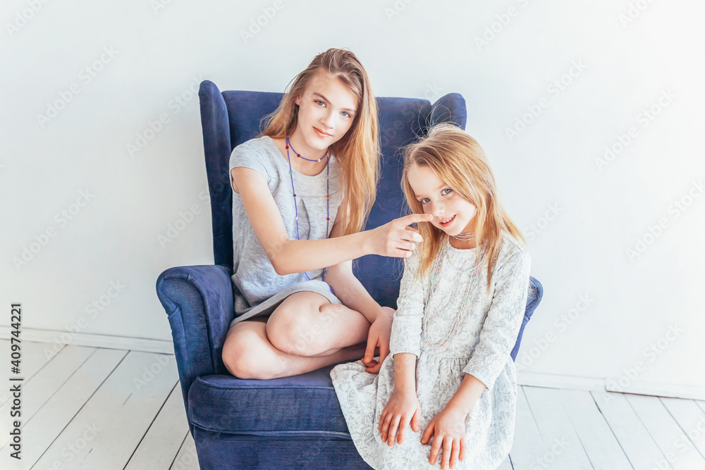Obraz premium Two happy kids sitting on cozy blue chair relaxing playing in white living room indoors. Little girl playing with teenage girl showing her love care. Sisters having fun at home
