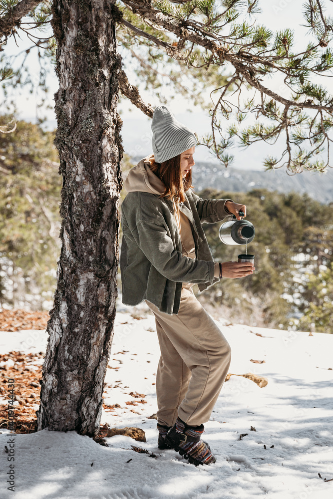 Girl in the mountains pours tea from a thermos. Winter and spring time. Dressed in a turquoise shirt and hat. Drink warm herbal tea in the cold. 